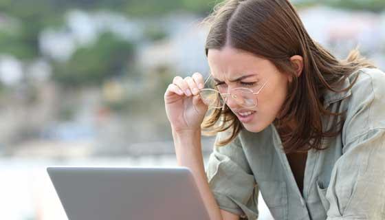 A young woman squints while lifting her glasses to see her laptop screen