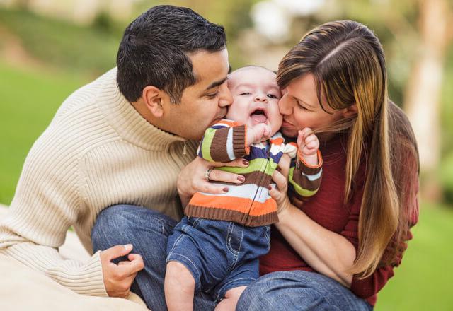 two parents kissing their infant on the cheek at the same time in a park